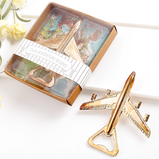 A gold-colored airplane-shaped bottle opener displayed next to its brown cardboard gift box with a clear window and a white paper band reading 'OUR ADVENTURE BEGINS!'. The scene is styled on a white surface with soft yellow flowers in the corner.